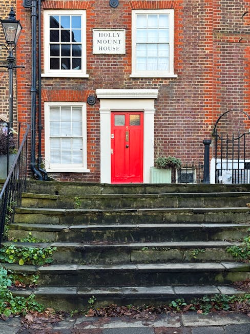 Exterior view of a red front door with a small window, set into a brick residential building named Holly Mount House, located in West Hampstead. The entrance is accessed via a set of weathered stone s